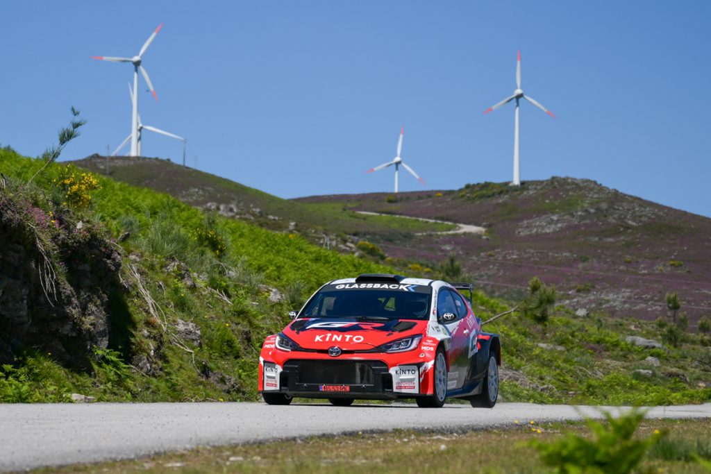 Veículo de corrida Toyota Gazoo Racing em movimento numa estrada rural com paisagem de montanhas e eólicas 