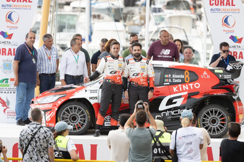 Dois pilotos de rally e um veículo de corrida da toyota em cima de um palco com pessoas à volta