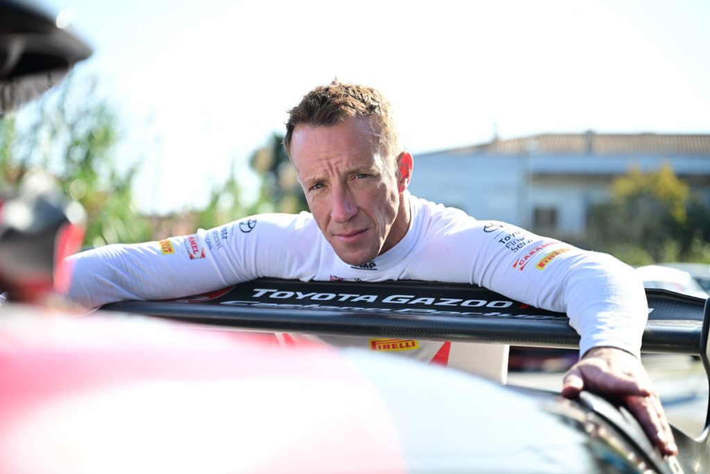 Kris Meeke, piloto de rali irlandês, fotografado de frente com um fato de competição da equipa TOYOTA GAZOO Racing Caetano Portugal e apoiado na parte traseira de um carro de competição Toyota.