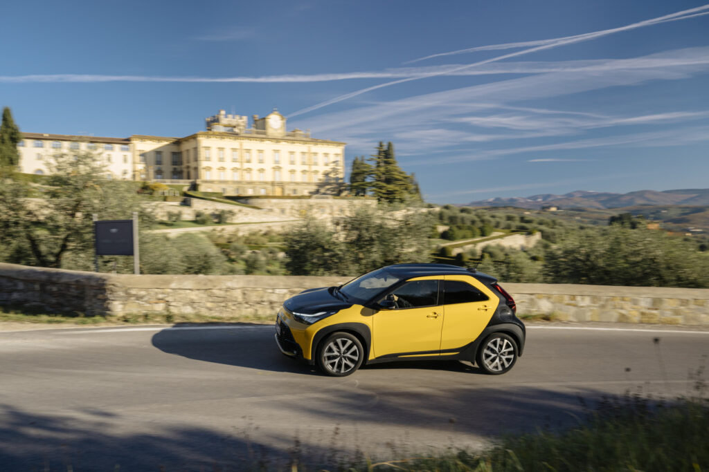 Toyota Aygo X Hybrid, na cor Mustard, exclusiva à versão GR Sport, em estrada com árvores e um edifício no fundo