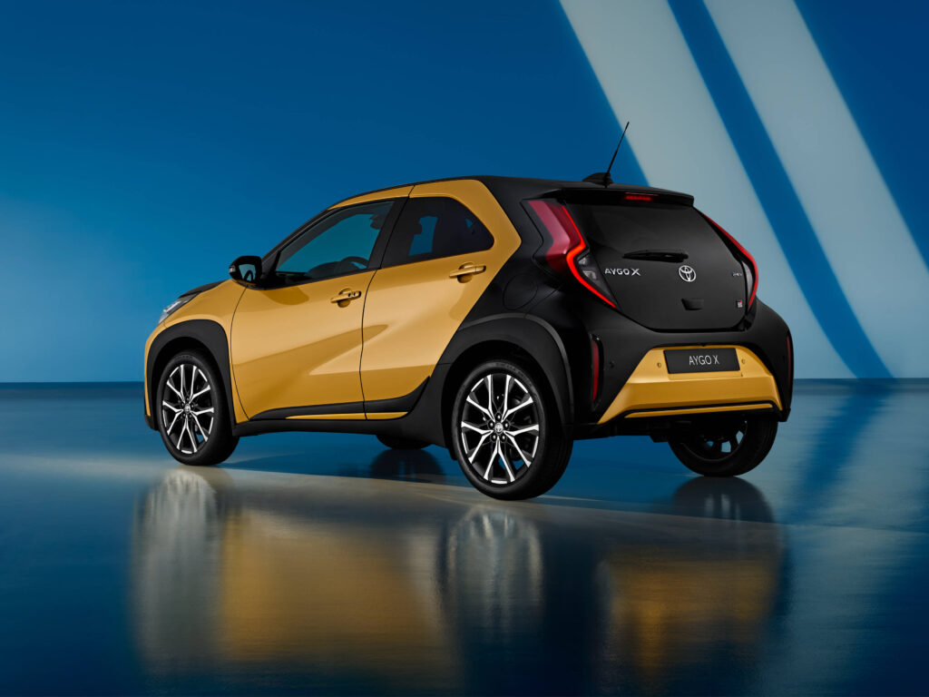 Vista 3/4 traseira do novo modelo Toyota Aygo X GR Sport na cor Mustard, com um fundo azul