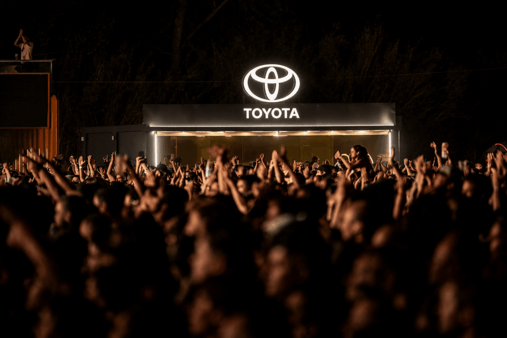 Conjunto de pessoas num festival, com os braços no ar, e stand da Toyota no fundo