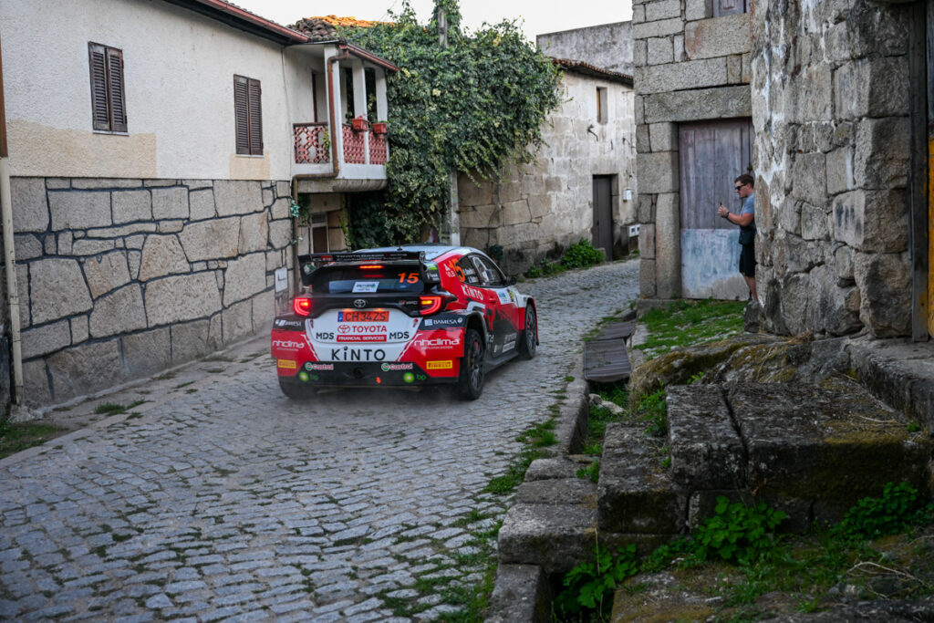 Fotografia da traseira do Toyota GR Yaris Rally2 pilotado por Pedro Almeida enquanto atravessa uma zona residencial no interior, espectador tira fotos ao automóvel de competição