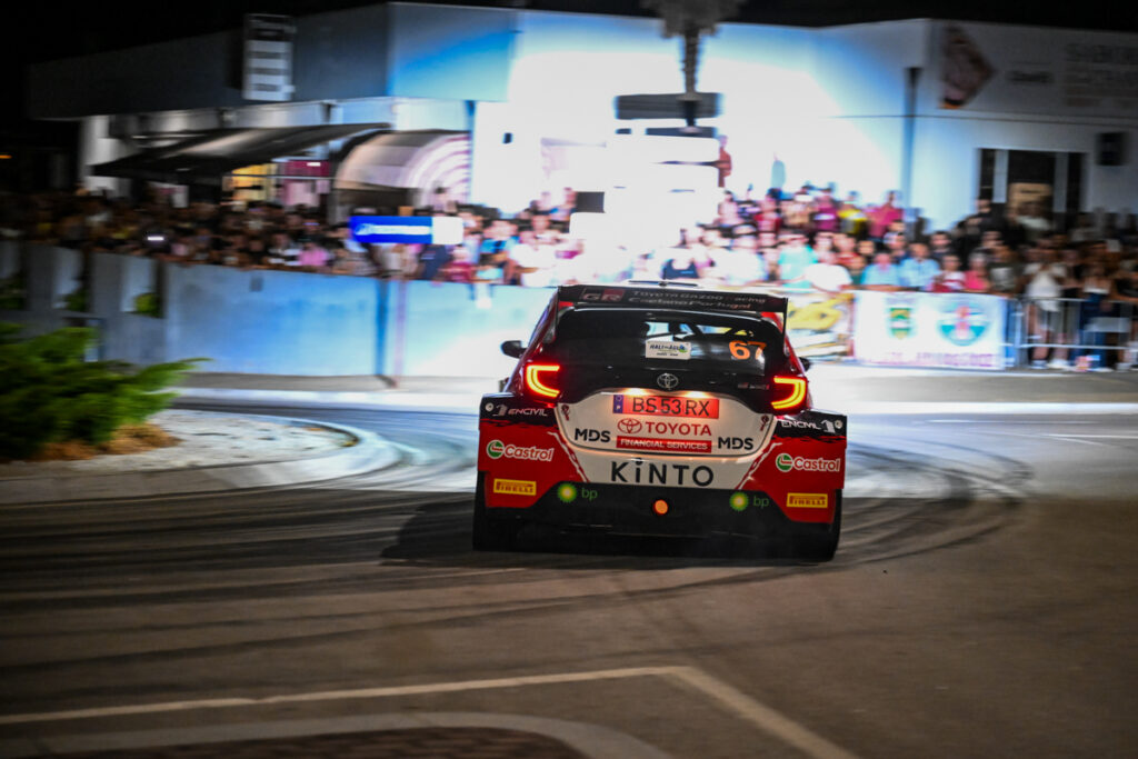 Traseira do automóvel de competição Toyota GR Yaris Rally2 do piloto Rafael Rêgo fotografado prestes a realizar uma curva em ambiente noturno com muitos espetadores no fundo